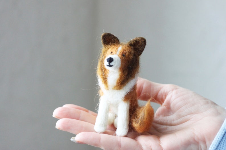 May include: A small, brown and white felt dog figurine with black eyes and a brown nose. The dog is sitting on a person's hand.