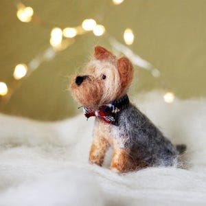 May include: A handcrafted Yorkshire Terrier dog figurine with a gray and tan coat, wearing a plaid bow tie. The miniature felted dog sits on a white, fluffy surface, with soft, blurred lights in the background.