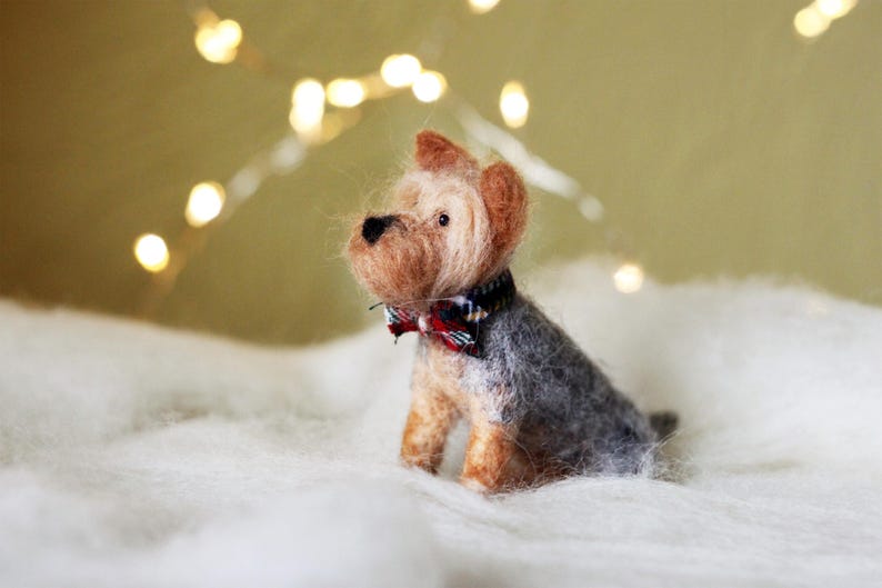 May include: A handcrafted Yorkshire Terrier dog figurine with a gray and tan coat, wearing a plaid bow tie. The miniature felted dog sits on a white, fluffy surface, with soft, blurred lights in the background.