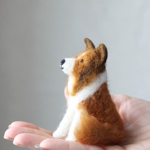 May include: A small, brown and white needle felted dog figurine. The dog is sitting up with its head tilted up and its ears perked.
