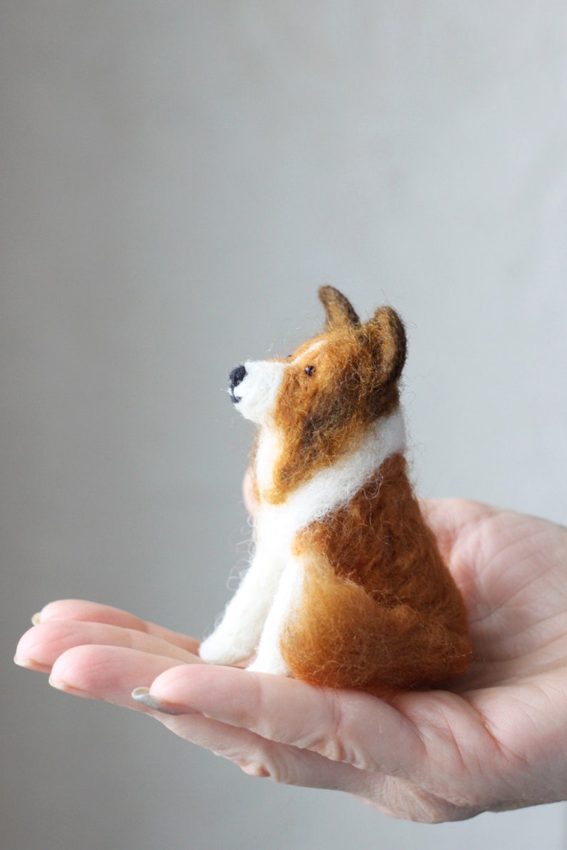 May include: A small, brown and white needle felted dog figurine. The dog is sitting up with its head tilted up and its ears perked.