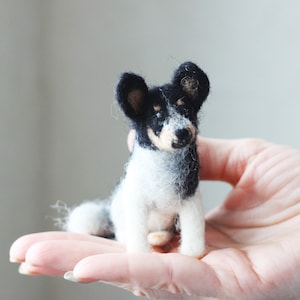 May include: A small, needle-felted dog figurine with black and white fur. The dog is sitting on a person's hand.