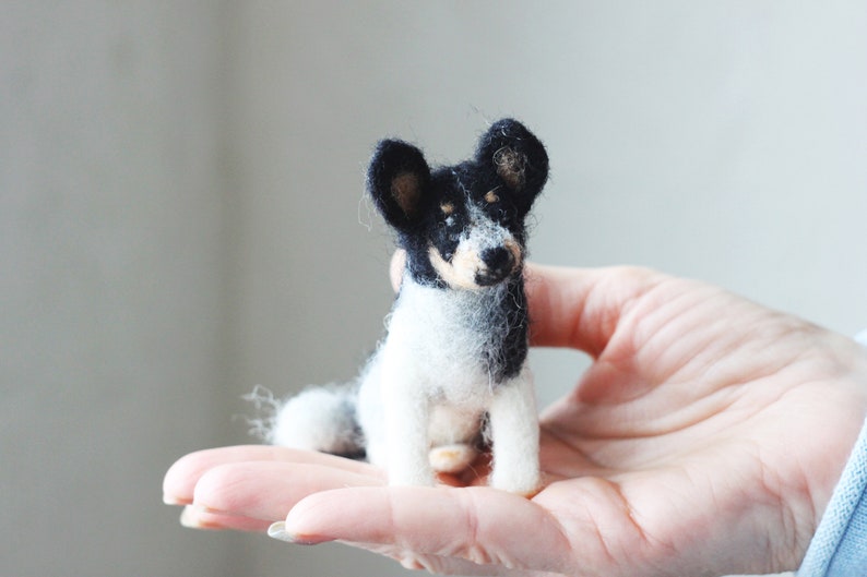 May include: A small, needle-felted dog figurine with black and white fur. The dog is sitting on a person's hand.