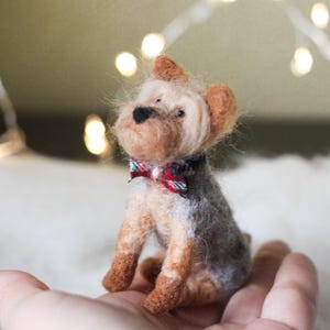May include: A handcrafted miniature Yorkshire Terrier dog figurine. The dog has a gray and tan body, with a red and blue plaid bow tie. The figurine is made of felted wool and is approximately 3 inches tall.