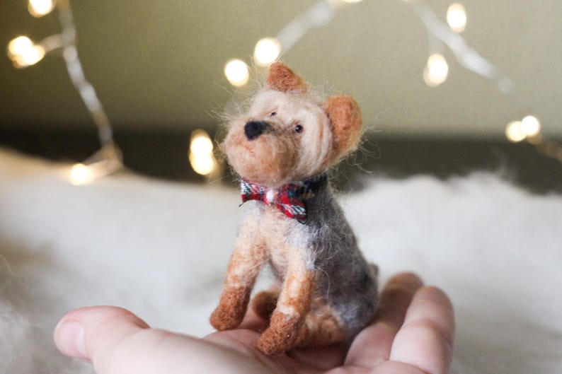 May include: A handcrafted miniature Yorkshire Terrier dog figurine. The dog has a gray and tan body, with a red and blue plaid bow tie. The figurine is made of felted wool and is approximately 3 inches tall.