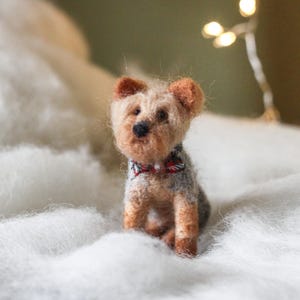 May include: A handcrafted Yorkshire Terrier dog figurine with a red, white, and black bow tie. The small dog is made of wool and sits on a bed of white fluff. The dog has brown and tan fur.