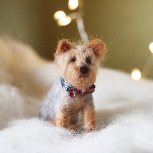 May include: A handcrafted miniature Yorkshire Terrier dog figurine. The dog has a light brown and gray coat, with a small blue and red bow tie. The figurine is sitting on a bed of white fluff, with soft lighting in the background.