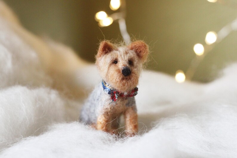 May include: A handcrafted miniature Yorkshire Terrier dog figurine. The dog has a light brown and gray coat, with a small blue and red bow tie. The figurine is sitting on a bed of white fluff, with soft lighting in the background.