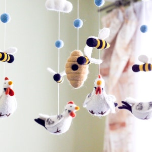 May include: A hanging mobile featuring felted wool figures suspended by white strings. The elements include white chickens with red combs, yellow and black striped bees, a tan beehive, a white cloud, and small light blue felted spheres.