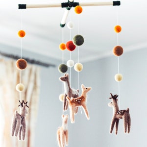 May include: A wooden mobile with felt deer and colorful pom poms hanging from it. The mobile is suspended from the ceiling.