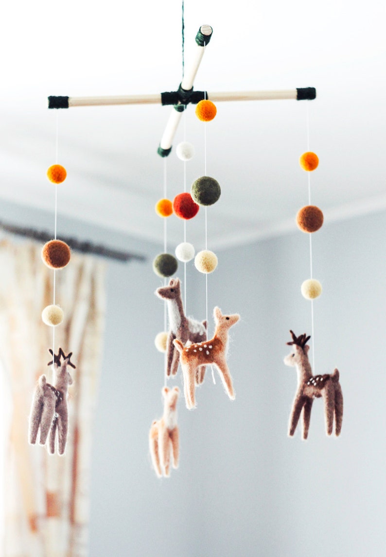 May include: A wooden mobile with felt deer and colorful pom poms hanging from it. The mobile is suspended from the ceiling.