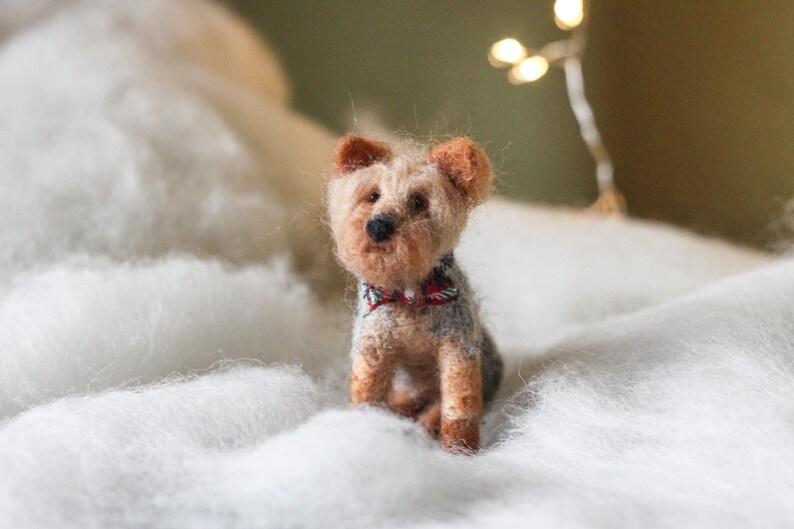 May include: A handcrafted Yorkshire Terrier dog figurine with a red, white, and black bow tie. The small, detailed dog is made of wool and sits on a bed of white fluff. The dog has brown and tan fur.
