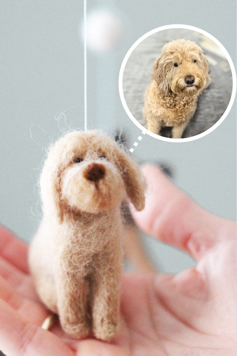 May include: A small, hand-felted dog figurine, made of light brown wool, with a fluffy texture. The figurine is sitting on a hand, with a thought bubble above it showing a larger, real dog of the same breed.