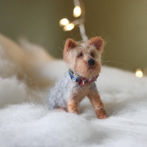 May include: A handcrafted Yorkshire Terrier dog figurine, made of wool, sits on a bed of white fluff. The dog has tan and gray fur, with a blue and red patterned collar. Soft, warm lighting and bokeh lights in the background.