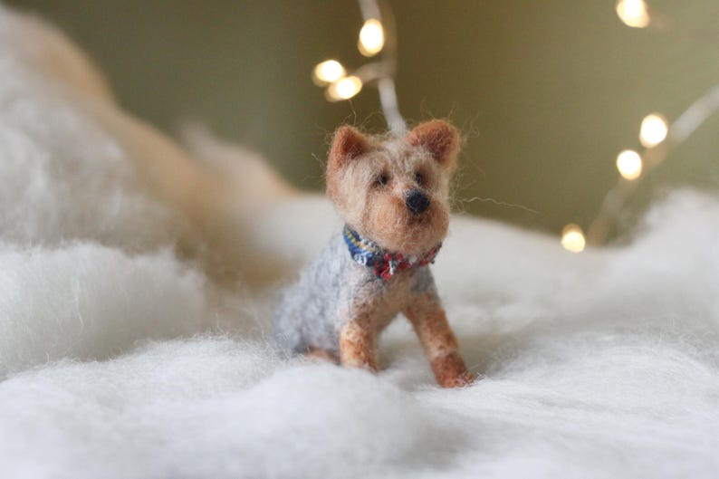 May include: A handcrafted Yorkshire Terrier dog figurine, made of wool, sits on a bed of white fluff. The dog has tan and gray fur, with a blue and red patterned collar. Soft, warm lighting and bokeh lights in the background.
