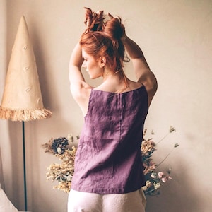 Könnte beinhalten: Eine Frau trägt ein lilafarbenes Tanktop aus Leinen und eine weiße Leinenhose. Sie steht vor einer Wand mit einem dekorativen kegelförmigen Objekt auf einem Ständer.