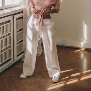 May include: A woman wearing a light pink wrap top with three-quarter sleeves and white wide-leg pants. The outfit is paired with white sneakers. The setting is a room with a window and wooden floor.