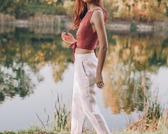 May include: A woman wearing a rust-colored crop top with a tie back and white linen pants. She is wearing brown espadrille flats. The background features a lake and trees.