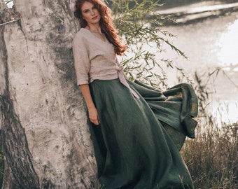 May include: A woman wearing a beige wrap top and a long, flowing, dark green skirt. The outfit is made of linen. The woman is leaning against a weathered tree trunk near a body of water. The scene is outdoors with natural light.