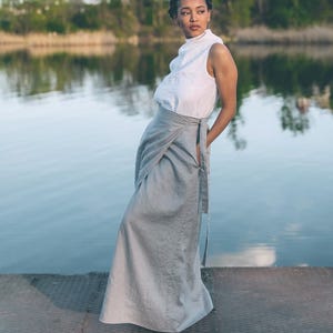 Può includere: Una donna indossa un top bianco senza maniche e una gonna a portafoglio grigio chiaro. La gonna lunga arriva fino a terra. Lo sfondo presenta acqua e alberi sotto una luce naturale soffusa.