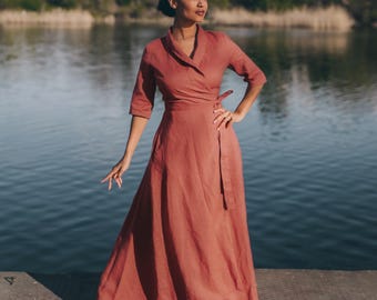 May include: A full-length, rust-colored wrap dress with three-quarter sleeves. The dress is made of a flowing fabric and is tied at the waist. The person is standing on a concrete surface near a body of water.