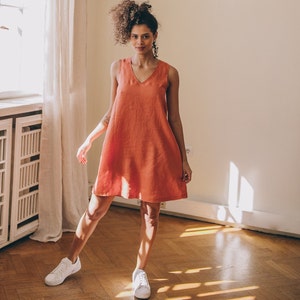 May include: A woman wearing a coral sleeveless linen dress and white trainers stands in a room with hardwood floors.