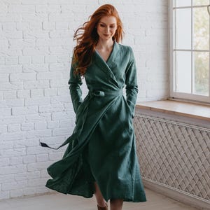 Könnte beinhalten: Ein langes, smaragdgrünes Wickelkleid mit langen Ärmeln und einem Taillenband. Das Kleid ist aus fließendem Stoff gefertigt und hat Taschen. Das Model trägt braune Stiefeletten. Der Hintergrund ist eine weiße Backsteinmauer und ein Fenster.