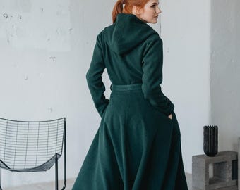 May include: A dark forest green hooded coat with a cinched waist, long sleeves, and a flared, knee-length skirt. The coat features a matching fabric belt at the waist and deep side pockets, worn over black leggings and black ankle boots.