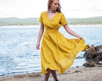 Op de afbeelding: Een vrouw draagt een gele wikkeljurk met korte mouwen op een strand. De jurk heeft een strik in de taille en valt tot halverwege de kuit. De achtergrond toont water en een blauwe lucht.