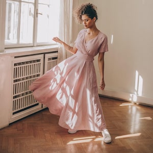 May include: A light pink wrap dress with short sleeves and a matching belt. The dress is made of a flowing fabric and is paired with white sneakers. The model is standing near a window with natural light.