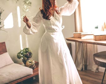 Puede incluir: Un vestido blanco cruzado con mangas largas y abullonadas. El vestido es largo hasta el suelo y se ata a la cintura. La imagen está tomada en una habitación con luz natural, un banco de madera y una alfombra.