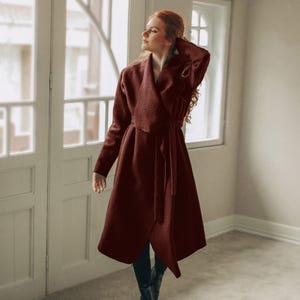 May include: A burgundy wrap coat with a matching belt. The coat has a shawl collar and long sleeves. The person in the image is wearing black boots. The coat is knee-length and made of a textured fabric.