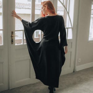 Puede incluir: Un vestido largo negro con mangas anchas y un gran corte circular en el lateral. El vestido lo lleva una persona con pelo largo y rojo, de pie cerca de una puerta blanca con ventanas arqueadas. El vestido parece estar hecho de una tela suave y fluida.