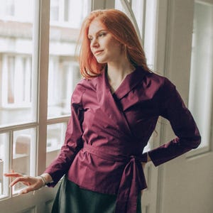 Könnte beinhalten: Ein weinrotes Wickeloberteil mit langen Ärmeln und passendem Bindegürtel, kombiniert mit einem dunkelgrünen Rock. Das Outfit ist aus glattem Stoff gefertigt. Die Oberteillänge beträgt ca. 60 cm.