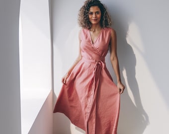 May include: A sleeveless, wrap-style dress in a soft coral color. The dress features a V-neckline and a matching fabric belt. The model is standing near a window, with natural light illuminating the dress and the white wall.