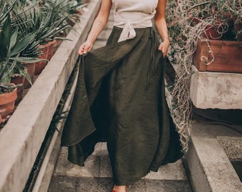 May include: A woman wearing a cream-colored top with a tie waist and a long, olive-green skirt. She is wearing red shoes. The setting is a greenhouse with various potted plants.