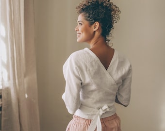 May include: White wrap top with a V-neck and short sleeves, paired with pink shorts. The top ties at the waist with a bow. The outfit is made of a lightweight fabric, perfect for warm weather. The clothing is casual and comfortable.