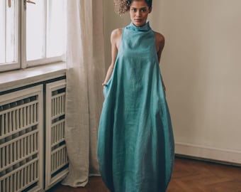 Puede incluir: Vestido sin mangas de color turquesa con cuello alto y corte amplio. La modelo está de pie en una habitación con ventana y suelo de madera. El vestido está hecho de una tela suave y ligera.