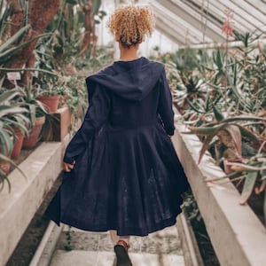May include: A person wearing a navy blue hooded coat with a flared skirt, walking through a greenhouse filled with plants. The coat has long sleeves and a hood. The person is wearing brown shoes.
