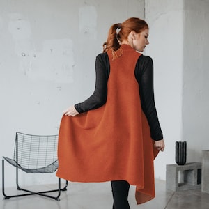 Peut inclure: Un gilet sans manches de couleur rouille avec un col montant. Le gilet est porté sur un haut noir à manches longues et des leggings noirs. La tenue est complétée par des bottines noires. Le gilet a un ourlet asymétrique.