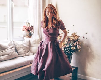 May include: A burgundy wrap dress with short sleeves and a flared skirt. The dress is cinched at the waist with a matching belt. The model is wearing brown sandals. The dress is made of linen. The setting is a well-lit room with a window and a round jute rug.