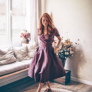 Könnte beinhalten: Burgunderfarbenes Wickelkleid mit kurzen Ärmeln und ausgestelltem Rock. Das Kleid ist mit einem passenden Gürtel in der Taille gerafft. Die Person trägt braune Sandalen. Das Kleid ist aus Leinen. Der Hintergrund ist ein gut beleuchteter Raum mit Fenster und rundem Jute-Teppich.