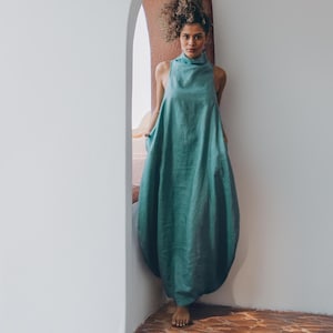 May include: A woman with curly hair wearing a teal linen dress stands in an arched doorway. The dress has a high neck and a loose, flowing fit.