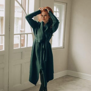 Puede incluir: Un abrigo cruzado de color verde oscuro con mangas largas, cuello ancho, cinturón para anudar a la cintura y dobladillo hasta la rodilla. El abrigo se lleva sobre unos leggings negros y botines negros, creando una silueta estructurada y fluida.