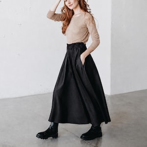 Pode incluir: Uma blusa bege de manga comprida com gola redonda combinada com uma saia longa preta de cintura subida com cós elástico e bolsos laterais, complementada por botins pretos com sola grossa e biqueira arredondada.