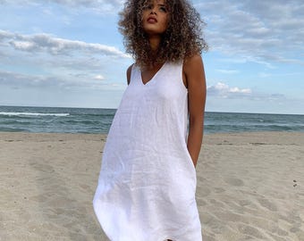 May include: A sleeveless white linen dress with a V-neck. The dress is knee-length and has pockets. The model is standing on a sandy beach with the ocean and a cloudy sky in the background.