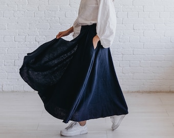May include: A woman wearing a white blouse with long, puffy sleeves and a long, navy blue skirt with pockets. The skirt is flowing, and the woman is wearing white sneakers. The background is a white brick wall.