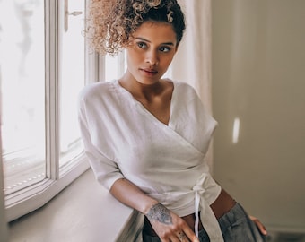 May include: A woman wearing a white wrap top with a tie closure and gray pants, leaning against a white window frame. The top has a V-neck and three-quarter sleeves. The woman has curly brown hair and a tattoo on her wrist.