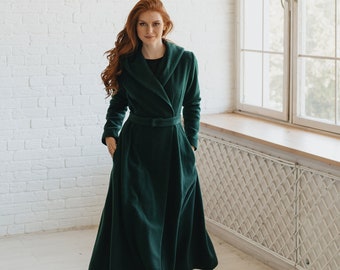 May include: A woman wearing a long, dark green coat with a hood and a belt. The coat has a wrap-style closure and a flowing skirt. She is standing in a room with white brick walls and a window.