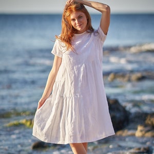 May include: A white linen dress with short sleeves and a tiered skirt. The dress is knee-length and worn by a person with red hair standing on a beach. The background features the ocean and a clear sky.